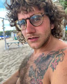 a shirtless man with glasses and tattoos on his chest standing in front of the beach