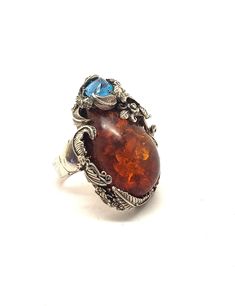 "What a stunning statement ring! Set with natural Amber and genuine Blue Topaz, this ring is truly a work of art. Handmade of sterling silver, the design features roses and flowing leaves. The Amber stone is large, about 35 mmm by 25 mm. The Blue Topaz is uniquely faceted. The whole setting is 1-3/4\" by 1-1/4\". The shank is a nice, heavy silver, 6 mm wide with a Claddagh design base. It appears to be signed, but unreadable. Unmarked, but guaranteed to be sterling silver." Handmade Topaz Rings With Unique Style, Unique Handmade Topaz Rings, Handmade Unique Topaz Rings, Unique Handmade Topaz Ring For Formal Occasions, Handmade Formal Topaz Ring, Unique Handmade Formal Topaz Ring, Unique Amber Wedding Rings, Natural Amber, Amber Stone