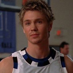 a young man wearing a white and blue basketball jersey looking at the camera while standing in a gym