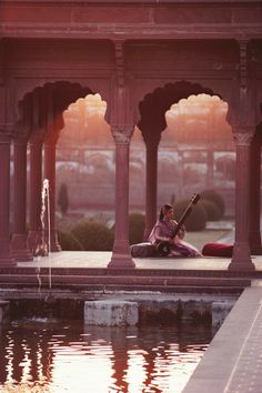 a woman sitting on the ground next to a body of water with an instrument in her hand