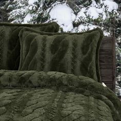 a green comforter and two pillows on a bed in front of snow covered trees