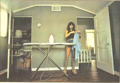 a woman ironing clothes on an ironing board in a room with gray walls