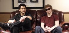 two young men sitting on brown leather couches in front of a framed photograph, one wearing sunglasses and the other with dark colored hair