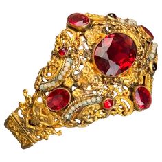 This exquisite bracelet, dating from the first half of the 20th century, showcases the remarkable craftsmanship of Austro-Hungarian jewelry. The wide band features vibrant, faceted garnet glass cabochons set in a luxurious gold gilt, accented by delicate faux seed pearls. Both the front and back of the bracelet display intricate openwork and pierced scroll designs, adding to its unique charm. The bracelet is equipped with a hidden, ornate tongue and box clasp, ensuring a secure fit while maintaining its elegant appearance. Although unmarked, the material is gold gilded metal, resembling lower grade silver or a metal mix, though it does not test positive for higher grade silver. Materials: Gold gilded metal Glass cabochons Faux seed pearls This stunning piece is perfect for collectors and t Austro Hungarian, Bracelet Display, Scroll Design, Gold Gilding, Wide Bands, Unique Charms, The 20th Century, Link Bracelets, A Metal