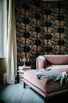 a pink couch sitting in front of a window next to a wall with flowers on it