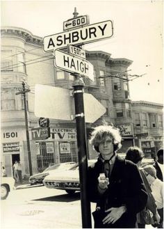 a woman standing next to a street sign on the side of a road with buildings in the background