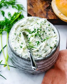 a bowl filled with white dip next to bread and green herbs on a cutting board