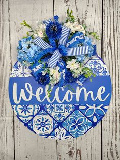 a welcome sign hanging on the side of a wooden wall with blue and white flowers