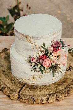 a three tiered cake with flowers on top sitting on a tree stump in the woods