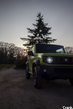 the front end of a green truck parked on a dirt road next to a pine tree