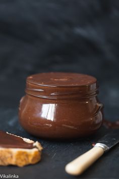 a jar of peanut butter next to a piece of bread on a black surface with a wooden spoon