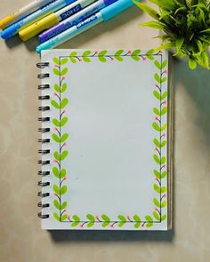 an open notebook with green leaves on it next to markers and a potted plant