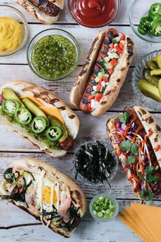 several hot dogs with toppings and condiments on a wooden table next to bowls of pickles