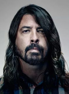 a man with long hair and beard wearing a plaid shirt looks at the camera while standing in front of a white background