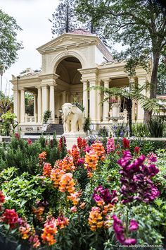 an elephant statue sitting in the middle of a garden with colorful flowers around it and a gazebo behind it