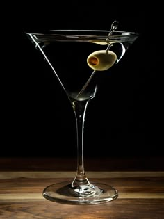 an olive in a martini glass sitting on a wooden table with a black back ground