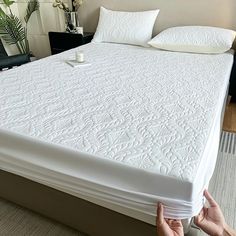 a person is holding up a mattress in front of a bed with white sheets and pillows