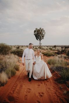 a bride and groom walking down a dirt path in the desert with a tree behind them