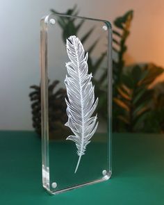 a clear glass block with a white feather etched on the front and back sides, sitting on a green surface