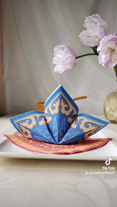 a blue and gold origami boat on a white plate with pink flowers in the background