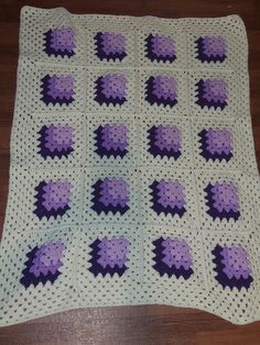 a crocheted blanket with purple and white squares on it sitting on top of a wooden floor