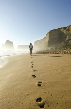 a woman walking on the beach with her footprints in the sand and cliffs in the background