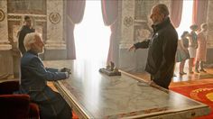 two men are sitting at a table in a room with red carpeting and large windows