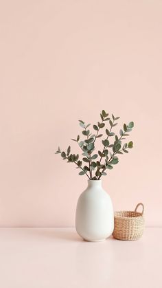 a white vase with green plants in it next to a basket on a pink surface