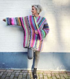 a woman leaning against a white brick wall wearing a multicolored knitted sweater