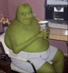 a large green creature sitting in front of a computer desk with a cup of coffee