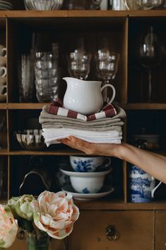 a person holding a stack of folded cloths in front of a wooden cabinet filled with dishes