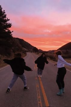 three young men skateboarding down an empty road at sunset with the sun setting in the background