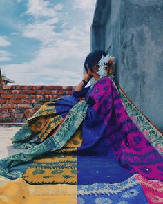 a woman sitting on the side of a building next to a pile of colorful blankets