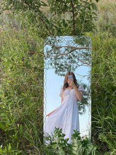 a woman taking a selfie in front of a mirror with trees and bushes behind her