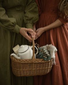 two women in dresses holding a wicker basket filled with tea pots and dishes,