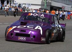 a purple race car driving down a track with people watching from the stands behind it