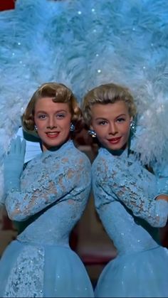 two women in blue dresses with white feathers on their head and arms, posing for the camera