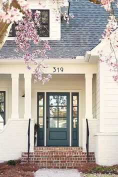 the front door of a white house with blue shutters and flowers in blooming trees