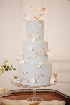 a three tiered blue cake with white flowers on the side and gold leaf decorations