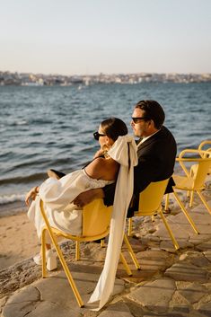 a man and woman sitting on yellow chairs by the water with their arms around each other