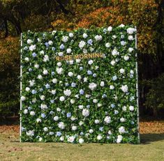 a green wall with white and blue flowers in the center is surrounded by trees that have leaves on them