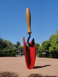 a large red sculpture with a wooden handle on it's head in the middle of a park