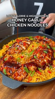 a pan filled with chicken and noodles on top of a wooden table next to a person