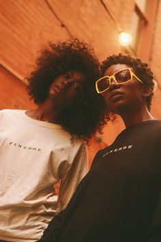 two young men standing next to each other in front of a brick wall wearing yellow sunglasses