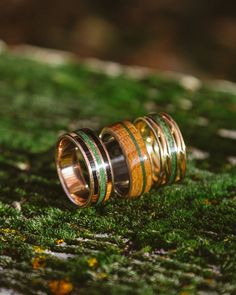 two wedding bands with green and yellow wood inlays are on top of a mossy surface