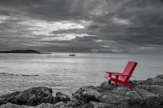 a red chair sitting on top of a rocky beach next to the ocean under a cloudy sky