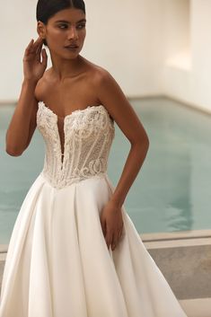 a woman in a white wedding dress standing next to a pool with her hand on her ear