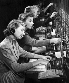 three women in headphones are working on an old fashioned computer screen with many rows of keyboards