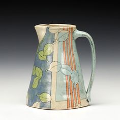 a ceramic pitcher with leaves and stripes on it's side, sitting on a table