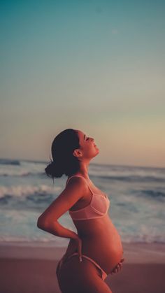 a pregnant woman standing on the beach looking up at the sky with her hands behind her back